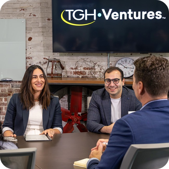 rachel feinman anthony zlaket and jason swoboda sitting at a conference room table in the tgh innovation center with a brick wall adorned with a monitor showing the tgh ventures logo in the background
