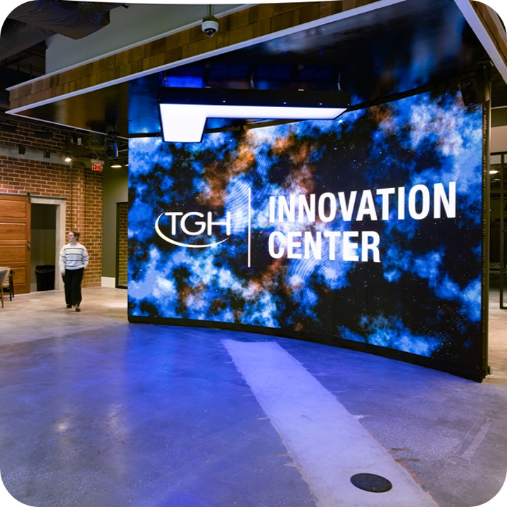a large led video wall showing the tgh innovation center logo and background graphic inside an industrial building with brick walls as a female walks by