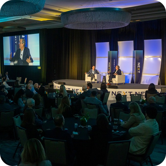john couris and jeb bush engaged in a fireside chat on stage at the next summit with a large crowd of attendees in the foreground